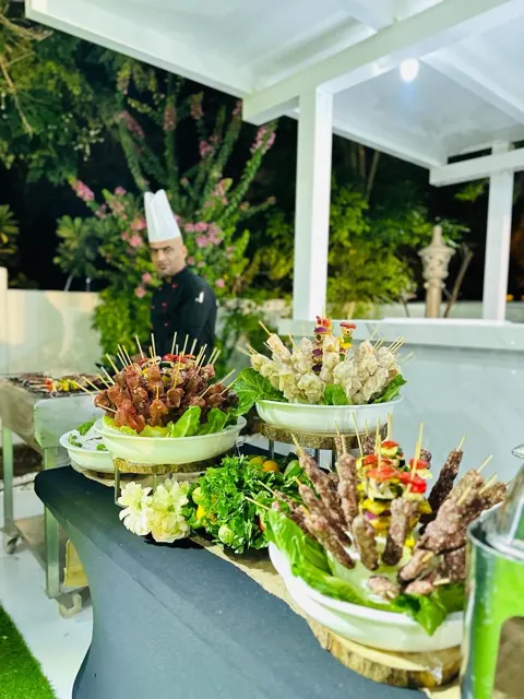 Chef at a garden BBQ station with skewers and vegetable displays.