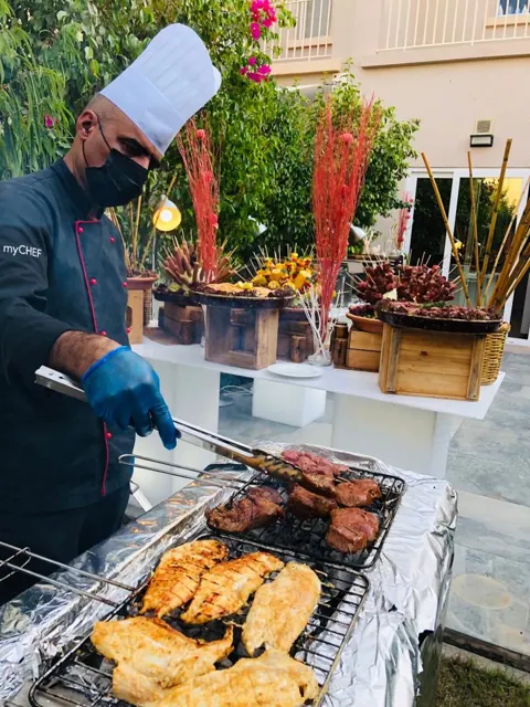 Chef turning food on an outdoor grill beside a villa.