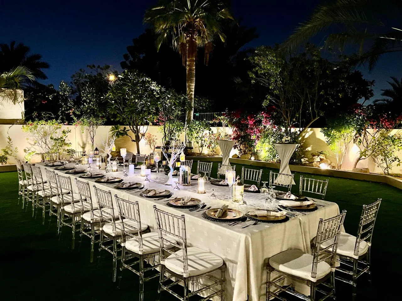 Long garden dinner table set with white chairs at night.