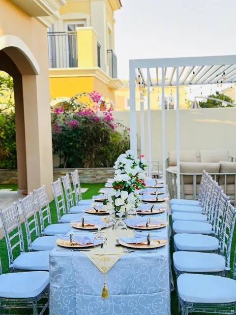 Long outdoor dining table with flowers and white chairs beside a villa.