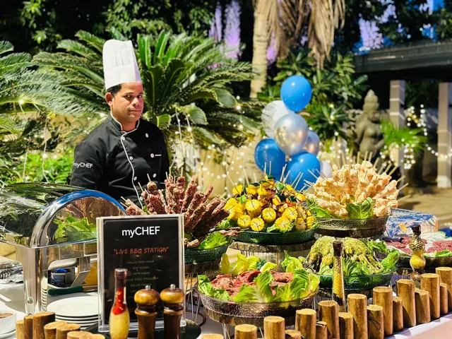Outdoor BBQ buffet with a chef, colourful skewers and blue balloons.