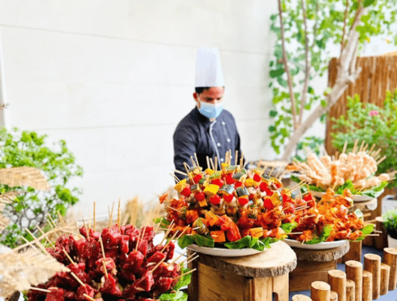 Outdoor BBQ skewers displayed beside a chef in a villa garden.